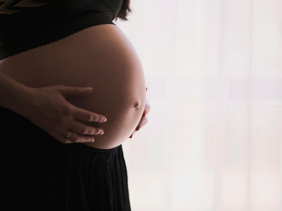 Mãos de uma gestante repousadas sobre a barriga, banhadas pela luz quente de uma janela ao fim de tarde.